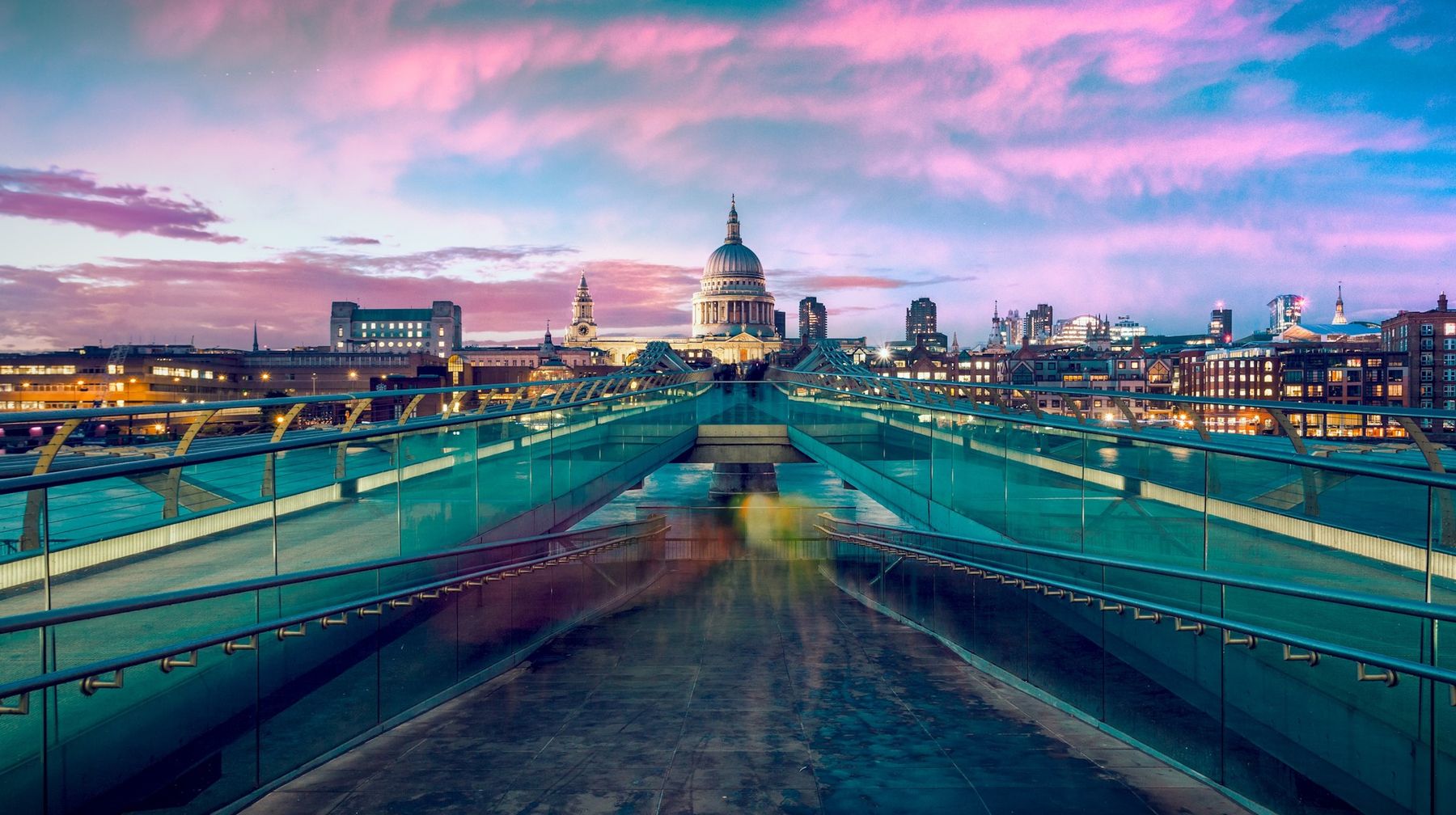 A footbridge over the Thames leading towards a domed cathedral under a pink dusk sky