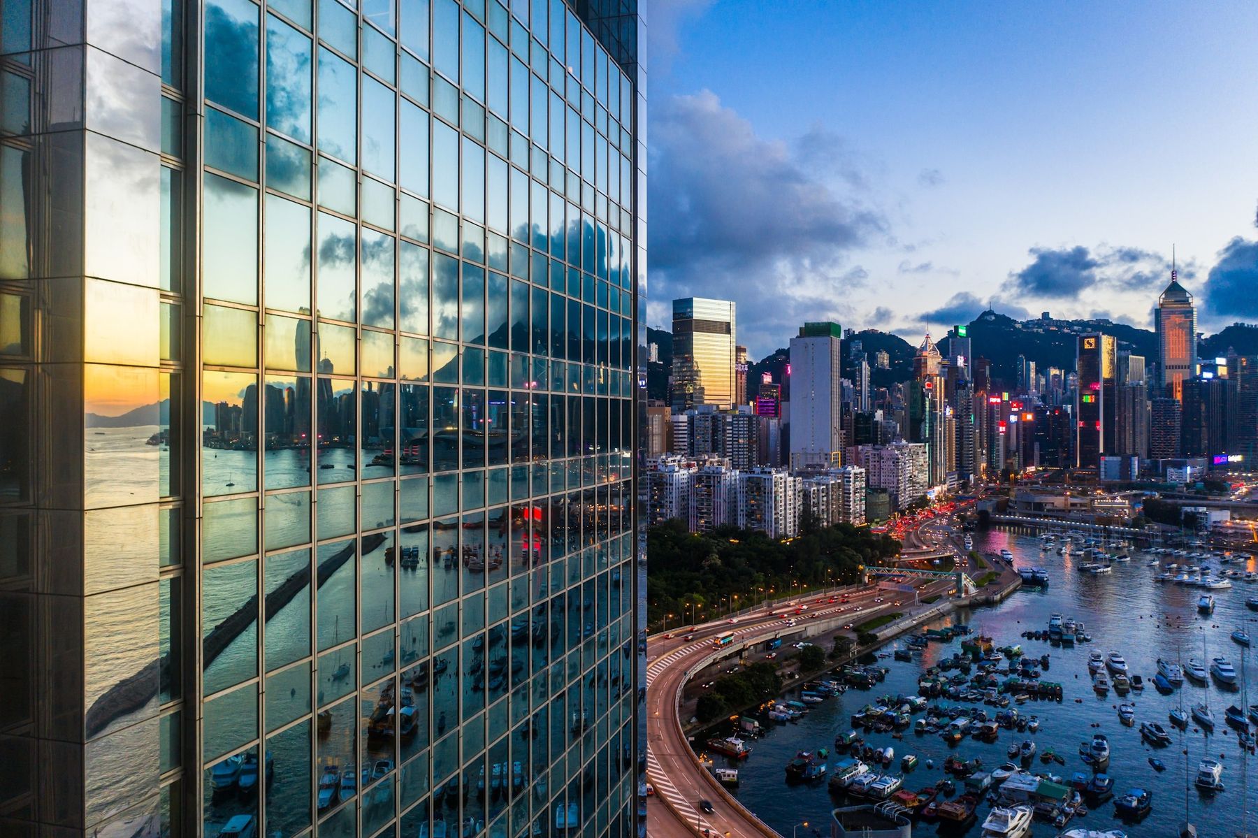 A glass office tower reflecting a city harbour at dusk, with high-rises and moored boats beyond