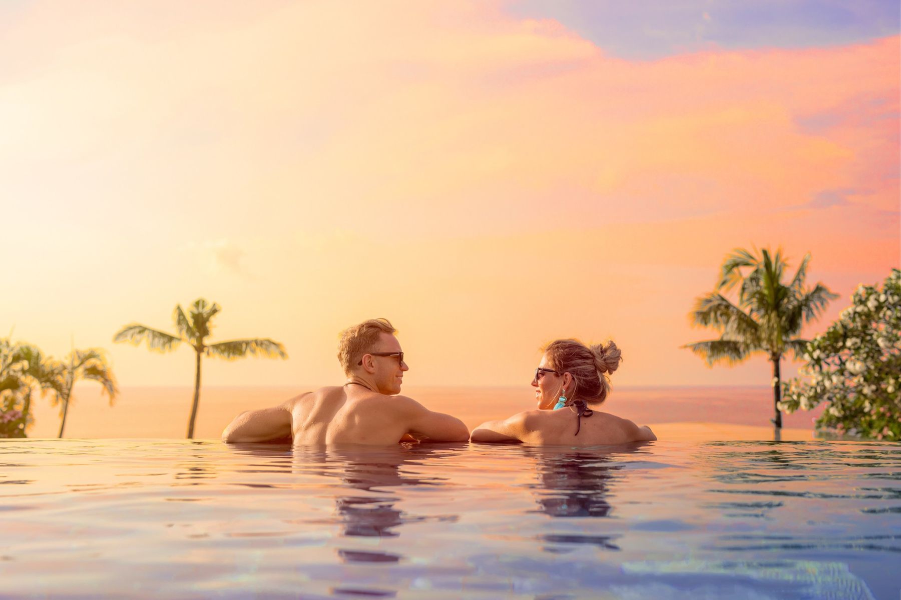 A couple relaxing at the edge of an infinity pool overlooking the sea at sunset, with palm trees on either side