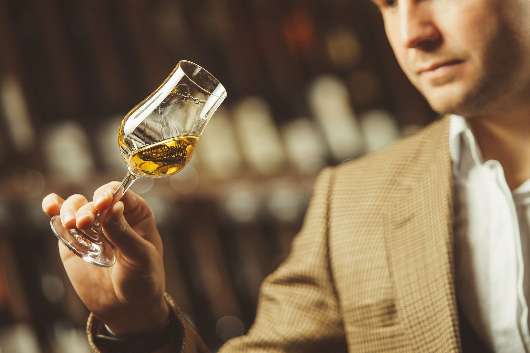 A man in a tweed jacket holding up a tulip glass of whisky in front of shelves of bottles